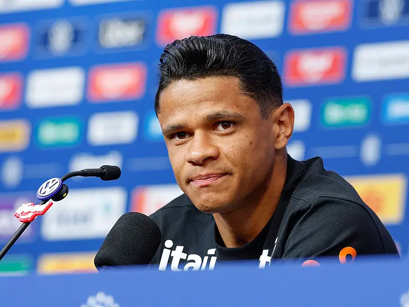 Jogador de futebol sorridente usando agasalho oficial de treino azul marinho, sentado em uma mesa de coletiva repleta de microfones