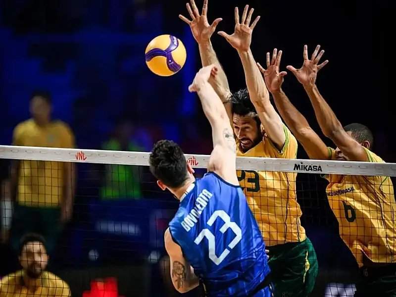 Jogadores da seleção de vôlei saltando na rede para tentar bloquear um ataque adversário em uma quadra oficial de vôlei iluminada