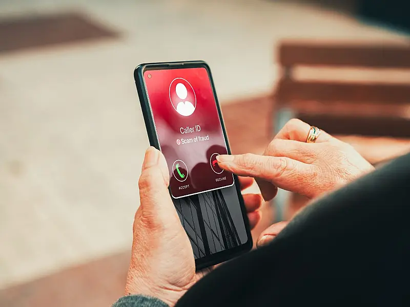 Mão de uma pessoa idosa segurando um telefone celular que exibe um alerta de chamada suspeita na tela de toque, com o dedo indicador prestes a recusar a ligação