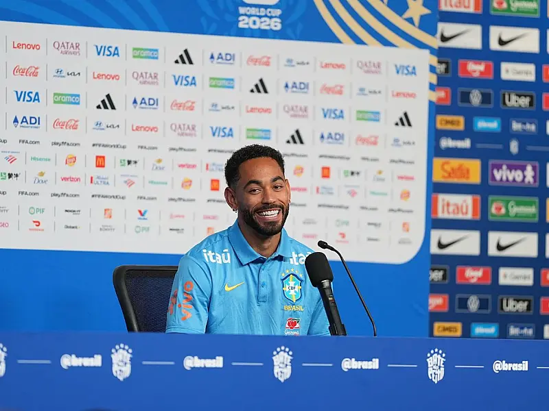 O atacante Matheus Cunha sentado à mesa de coletiva de imprensa com microfone à frente, vestindo o agasalho oficial azul da Seleção Brasileira de futebol