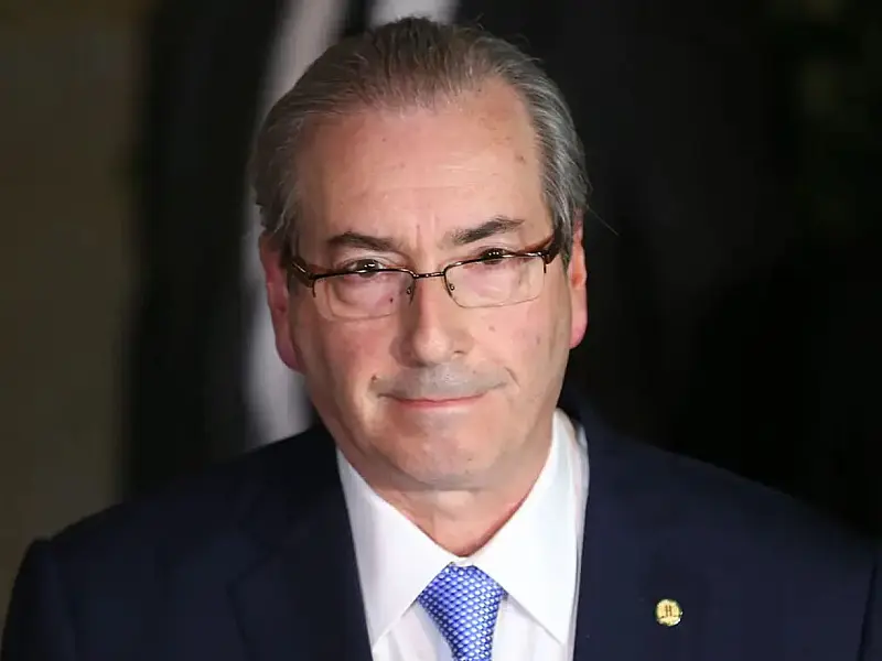 Retrato em plano médio de Eduardo Cunha de terno e gravata, com expressão séria, em um ambiente público iluminado