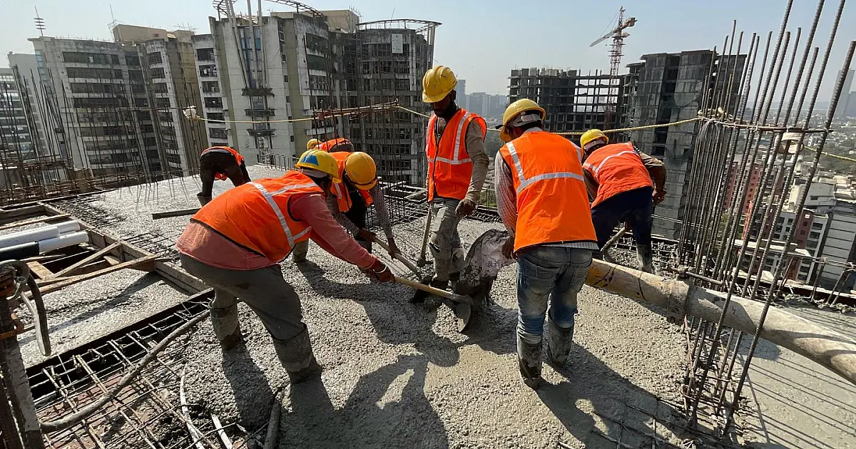 Fotografia em plano médio de operários com capacetes de segurança e coletes reflexivos trabalhando estruturalmente no topo de um arranha-céu em construção, contra um céu claro