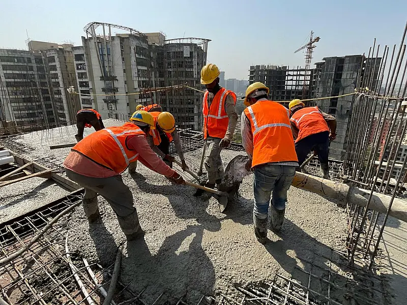 Fotografia em plano médio de operários com capacetes de segurança e coletes reflexivos trabalhando estruturalmente no topo de um arranha-céu em construção, contra um céu claro