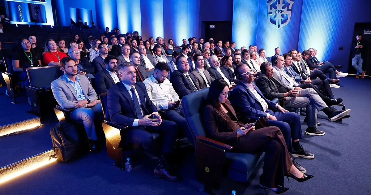 Sala de conferências da CBF com dirigentes e presidentes de federações estaduais sentados ao redor de uma grande mesa de reuniões, prestando atenção à apresentação projetada em uma tela ao fundo