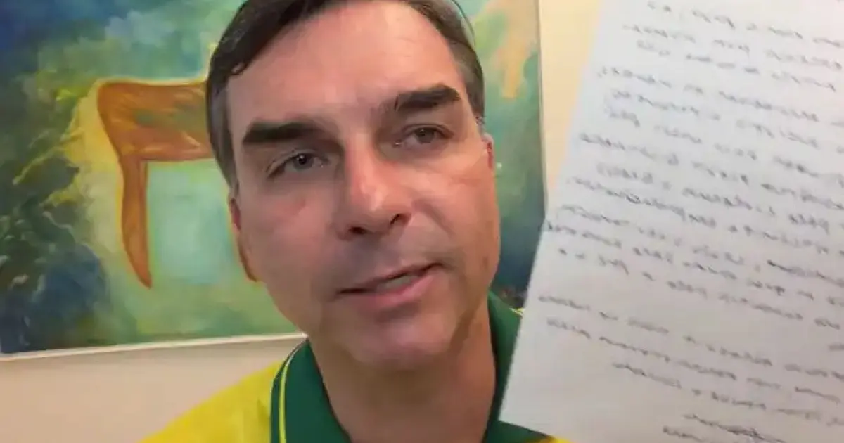 Fotografia do senador Flávio Bolsonaro em um ambiente interno, segurando folhas de papel contendo o manifesto escrito de seu pai, durante pronunciamento político
