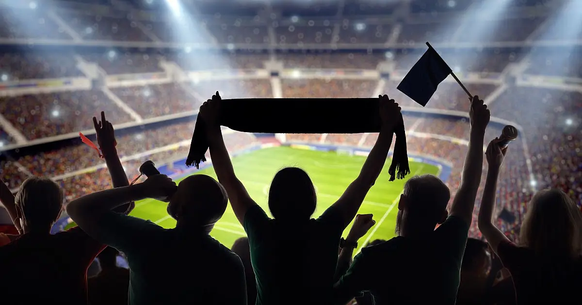 Vista geral das arquibancadas de um estádio de futebol lotado, com torcedores vibrando sob a luz dos refletores em uma noite de partida
