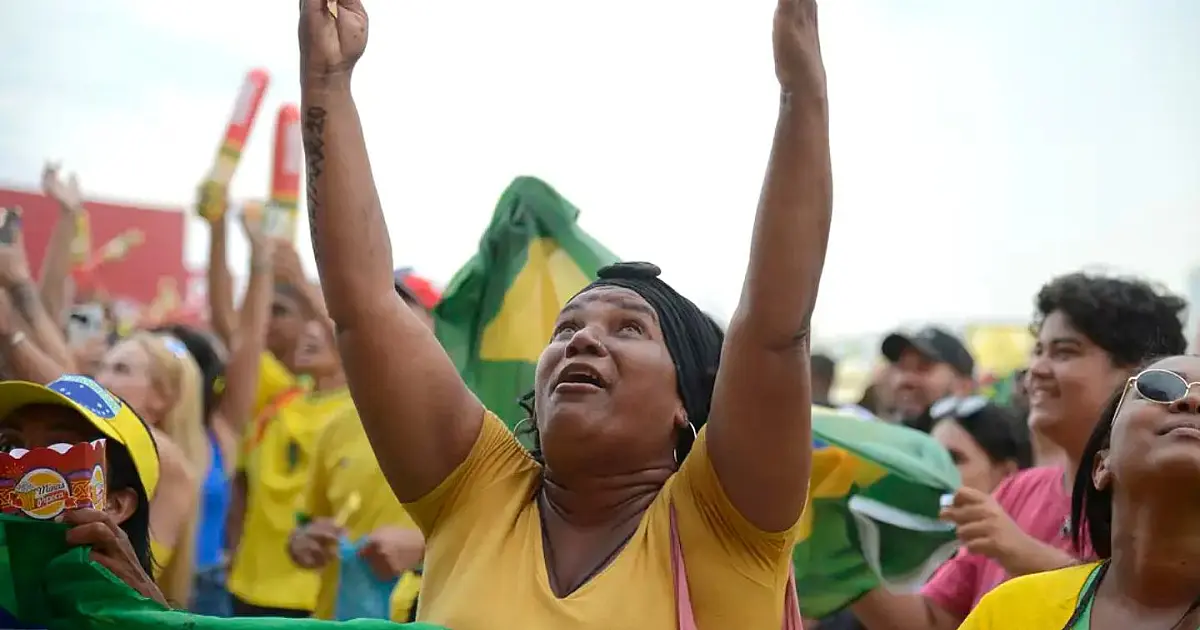 Vista de costas de uma multidão de torcedores vestindo camisas amarelas do Brasil celebrando em uma rua enfeitada com fitas coloridas