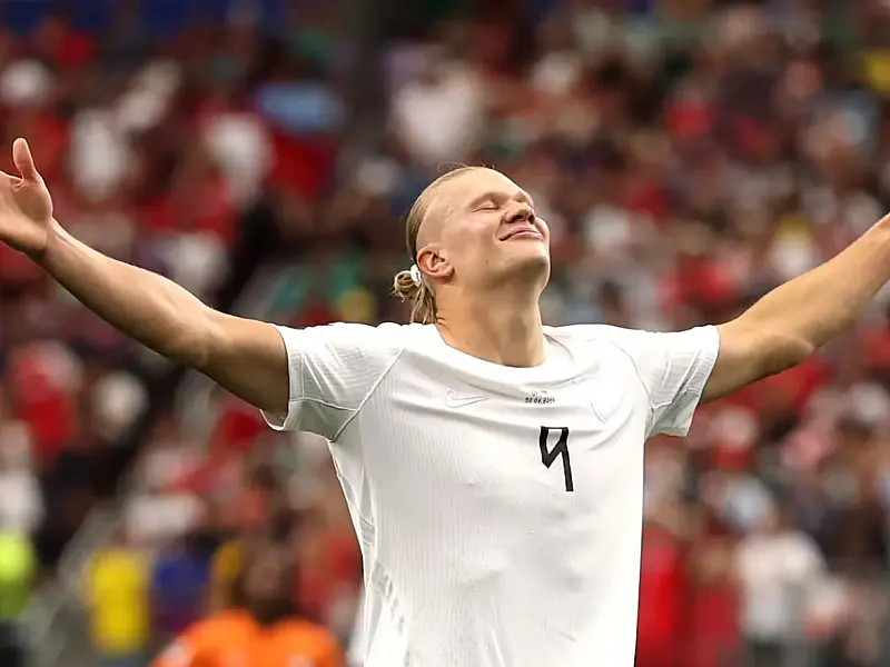 O atacante Erling Haaland comemorando o gol da vitória, correndo pelo gramado com os braços abertos e vestindo o uniforme vermelho da seleção da Noruega