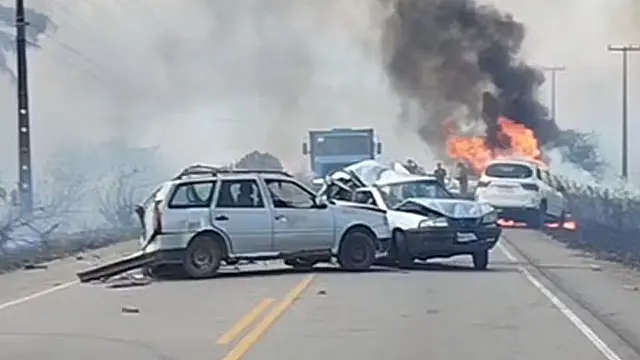 Carros destruídos por colisão e fogo em trecho de rodovia coberto por fumaça.